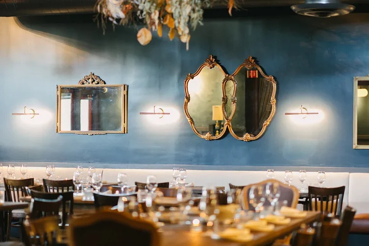 Elegant dining room with a blue wall decorated with vintage mirrors and modern wall sconces, set with wine glasses and tableware.