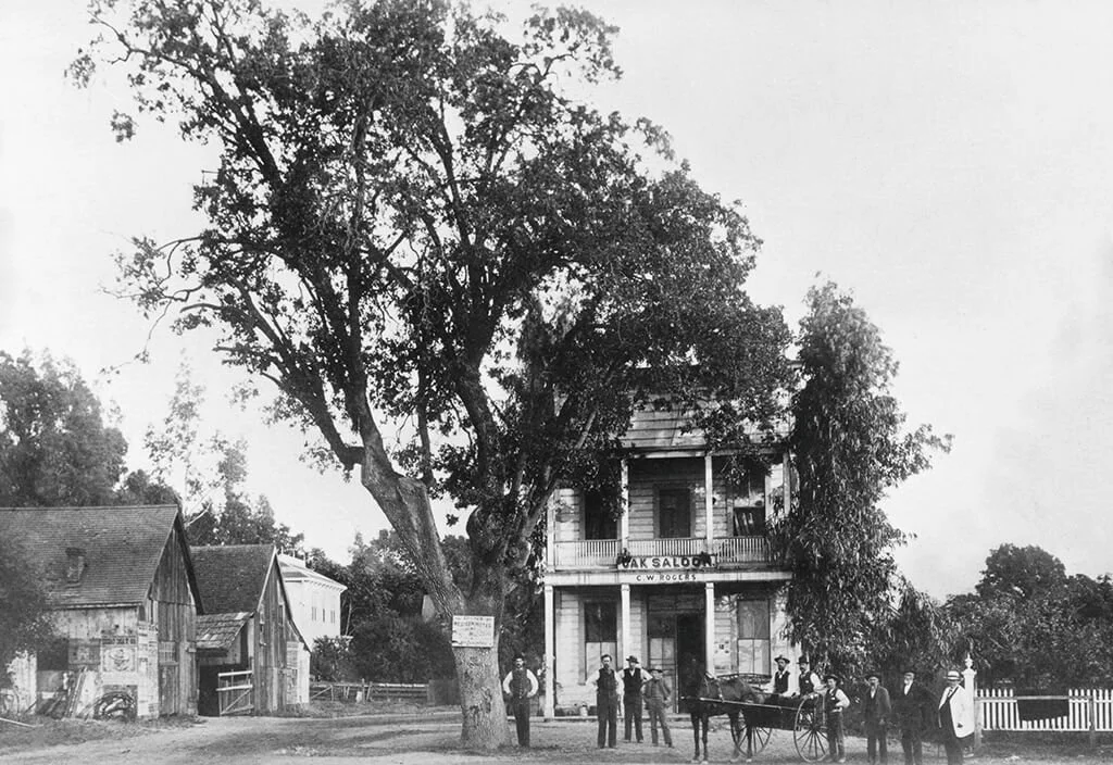 Historical black and white photo of a two-story building labeled 'Oak Saloon' with a porch, surrounded by trees and smaller wooden buildings, with several men standing outside and a horse-drawn cart in front.