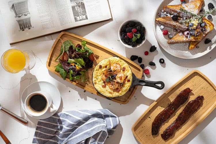 Overhead view of a breakfast spread with scrambled eggs, French toast with berries, orange juice, coffee, and glazed ribs.