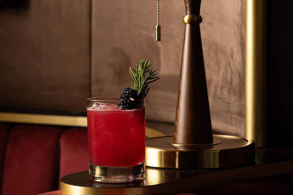 Deep red blackberry cocktail beside a brass lamp on the velvet banquette